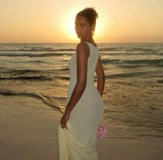 a woman standing on the beach at sunset wearing a white dress and holding a pink flower in her hand