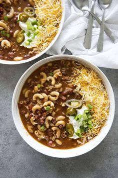 two bowls of chili and bean soup on a gray surface with spoons next to it