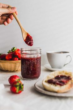 someone holding a spoon full of strawberry jam next to a bowl of strawberries and bread