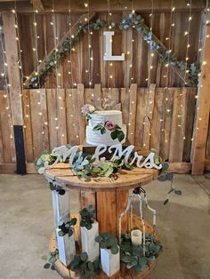 a wedding cake on top of a wooden table with greenery and lights around it