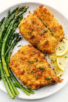 two pieces of chicken with asparagus on a white plate next to lemon wedges