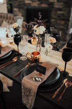 the table is set with black plates and silverware, gold place settings and white flowers