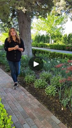 a woman walking down a brick path next to a tree and flowers on the ground