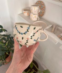 a hand holding up a coffee cup in front of a shelf with dishes on it