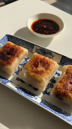 three pieces of food on a blue and white plate next to a bowl of dipping sauce