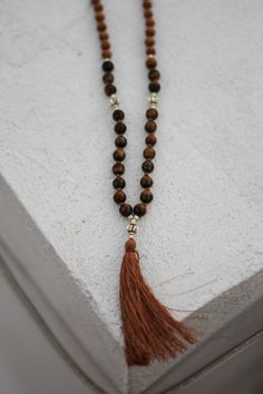 this 108 bead mala is designed by here & there creative and handmade in northern india with sandalwood and lava stone beads. the beads originate from the spiritual lands of northern india and have been blessed by the holy ganges river.  the tiger's eye beads vary in color from golden yellow to deep red. tiger's eye is a protection agent known as the "all-seeing all-knowing eye." with this mala, you'll have an added sense of security and comfort. here & there styling tip: if the tassel strands be Spiritual Healing Brown Beads, Brown Spiritual Beads For Meditation, Bohemian Brown Mala With Natural Stones, Hand-strung Brown Mala For Healing, Brown Mala With Round Beads For Healing, Brown Bohemian Mala With 108 Beads, Spiritual Brown Gemstone Beads, Bohemian Brown Mala With 108 Beads, Spiritual Brown Mala For Meditation