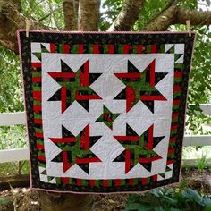 a quilt hanging from a tree in front of a white fence with green and red stars on it