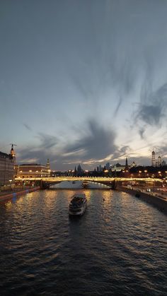 a boat traveling down a river next to a bridge and buildings at night with clouds in the sky