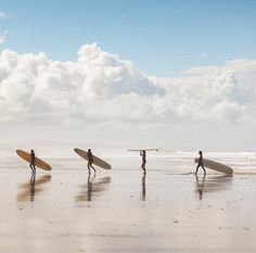 four people with surfboards are walking on the beach near the water and clouds in the sky