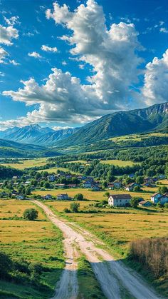 a dirt road in the middle of a field with houses and mountains in the background