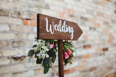 a wooden sign that says wedding with flowers and greenery attached to it, standing in front of a brick wall