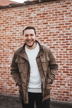 a man standing in front of a brick wall wearing a brown jacket and white sweater