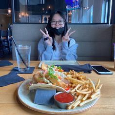 a woman sitting at a table with a plate of food in front of her, making the peace sign