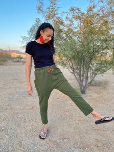 a woman in black shirt and khaki pants standing on dirt ground with trees behind her