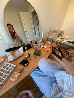 a woman sitting in front of a mirror looking at her cell phone and makeup products