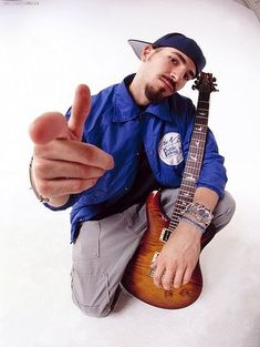 a man sitting on the ground holding a guitar and giving a thumbs up sign with his right hand