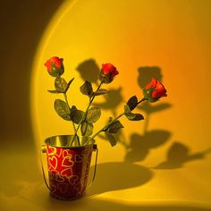 a red vase with flowers in it sitting on a yellow table top next to a shadow cast wall