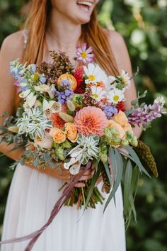 a woman holding a bouquet of flowers in her hands and smiling at the camera while wearing a white dress