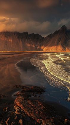 the sun is setting on an ocean beach with mountains in the background and dark clouds