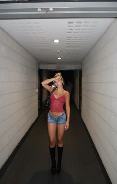 a woman in short shorts and boots is standing in an empty hallway with her hand on her head