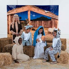 three children dressed up as nativity characters sitting on hay bales in front of a christmas scene