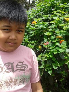 a young boy is standing in front of some bushes and flowers with his head turned to the side