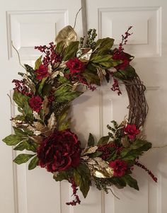 a wreath with red flowers and greenery hangs on a door handle, in front of a white door