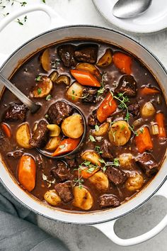 a pot filled with stew and carrots on top of a white table next to a spoon