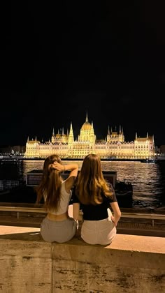 two women are sitting on a ledge looking at the water and buildings in the background