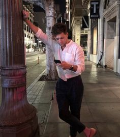 a man standing next to a pole on the side of a street at night looking at his cell phone