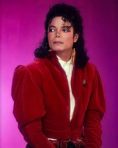 a man with long black hair wearing a red jacket and white shirt in front of a purple background