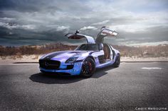 a blue sports car with its doors open sitting on the side of the road in front of a cloudy sky