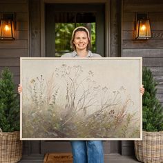 a woman holding up a large painting in front of a house with potted plants