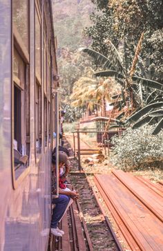 two people sitting on the side of a train as it passes by some trees and bushes