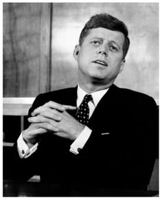 a black and white photo of a man in a suit sitting at a desk with his hands folded