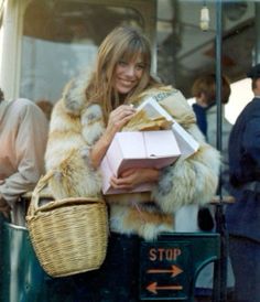 a woman in fur coat holding shopping bags and smiling at the camera while standing next to a bus