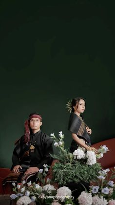 two people sitting on a couch with flowers in the foreground and a green wall behind them