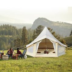 four people sitting in chairs around a tent on top of a grass covered field with mountains in the background