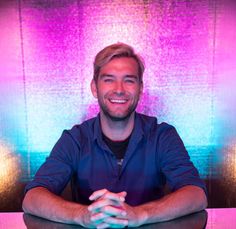 a man sitting at a table in front of a purple and blue background with his arms crossed