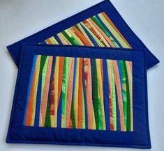 two blue place mats with strips of colored fabric on top of each other in front of a white background