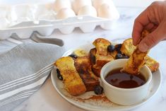 someone dipping some blueberries into a cup of sauce on a plate with french toast