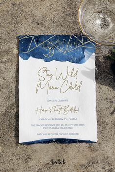 a blue and white wedding card on the sand next to a wine glass with water in it