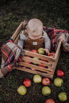 a baby sitting in a wooden crate with apples on the ground next to it's legs