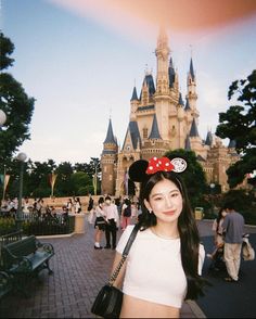 a woman standing in front of a castle with minnie mouse ears on her head and people walking around