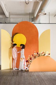 two people standing next to each other in front of an orange and yellow wall with flowers