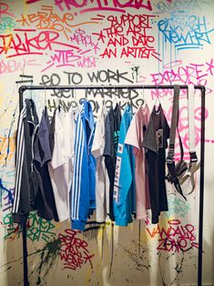 several shirts hanging on a rack in front of a wall with spray painted words and graffiti