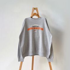 a gray sweatshirt hanging on a wooden hanger next to a white wall with an orange and black bronco's logo