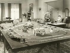 an old black and white photo of two men in a living room with a model train set on the table