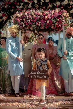 a group of people standing around each other in front of a flower covered arch with a sign