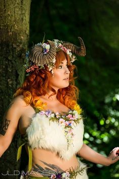 a woman with horns and flowers on her head is standing in front of a tree